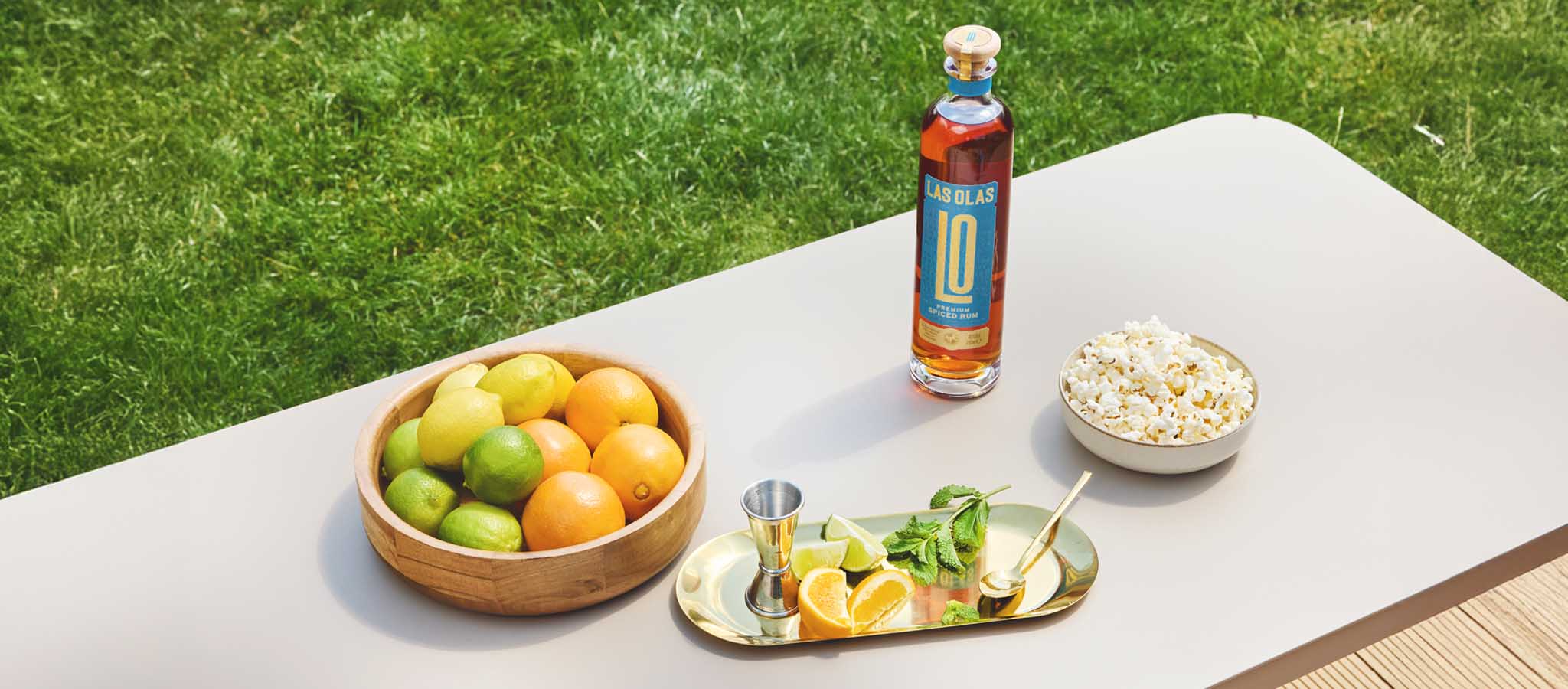 Bottle of Las olas with two cocktails on a tray against a with citrus fruits in one bowl and popcorn in another set against green grass