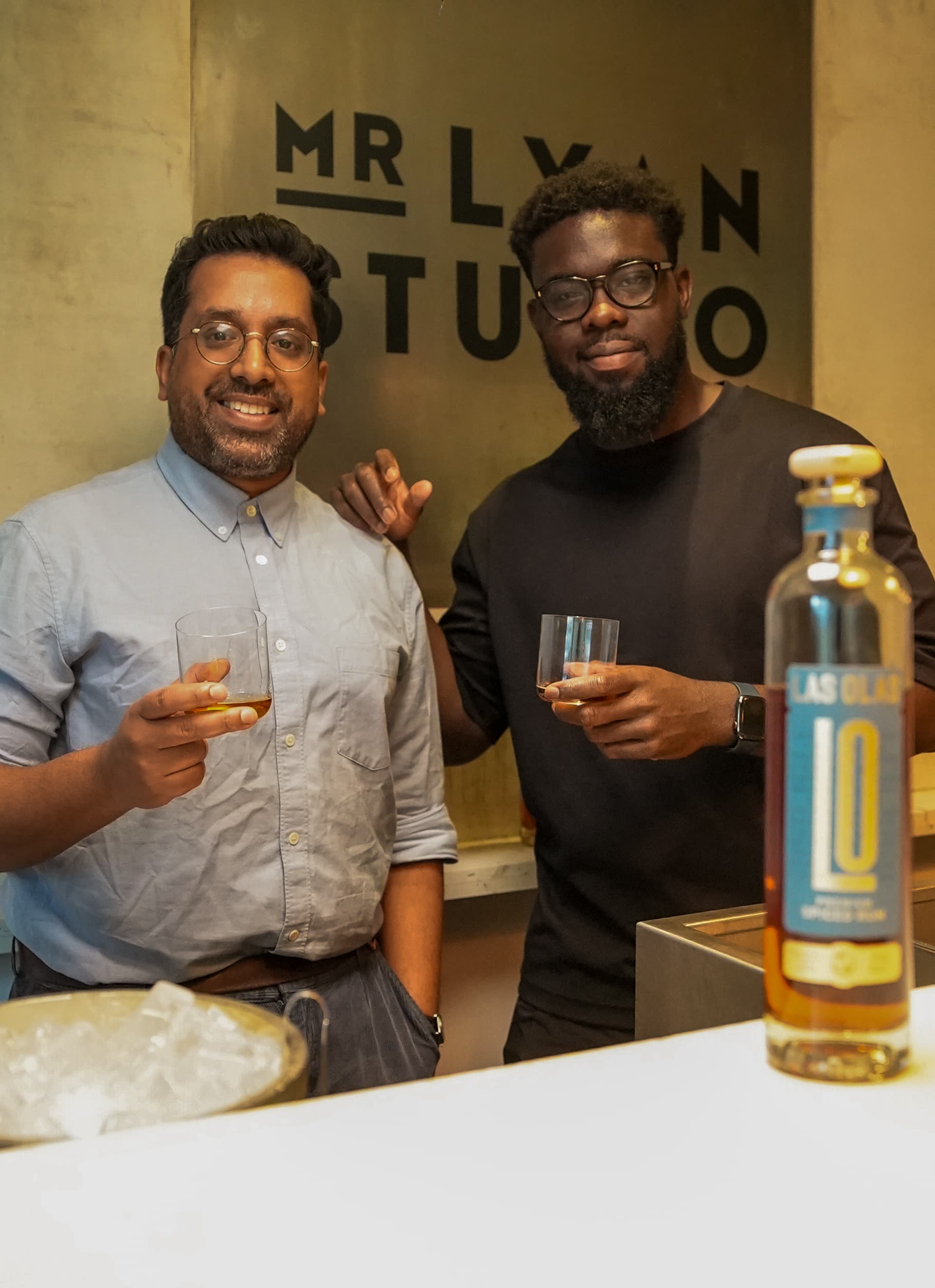 Two men holding drinks in front of a bottle with 'Mr Lyon Studio' in the background