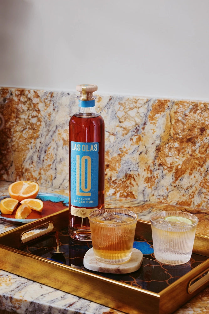 Bottle of Las Olas rum on a tray with two glasses filled with a yellowish liquid, against a marble wall.