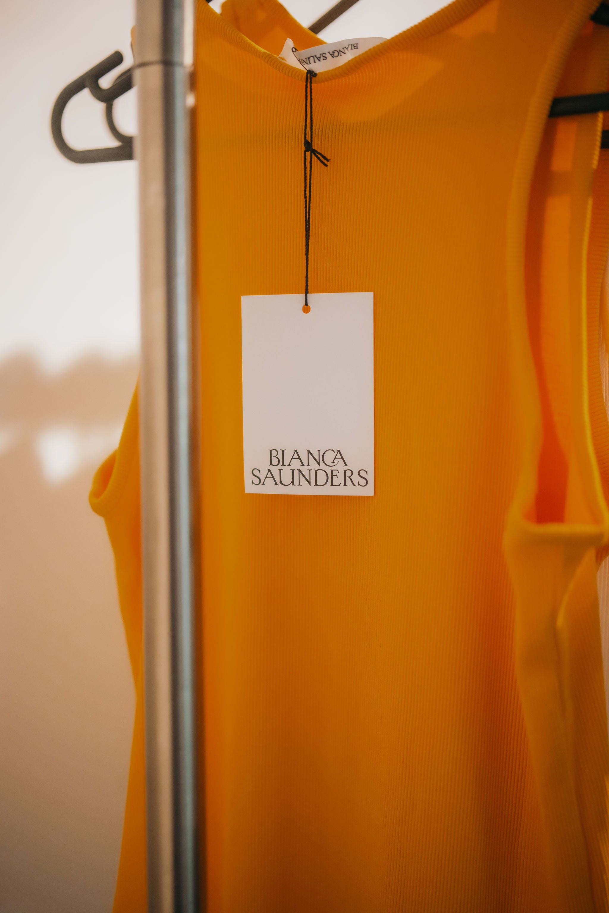 Yellow hoodie with a 'Bianca Saunders' tag on a hanger against a blurred background