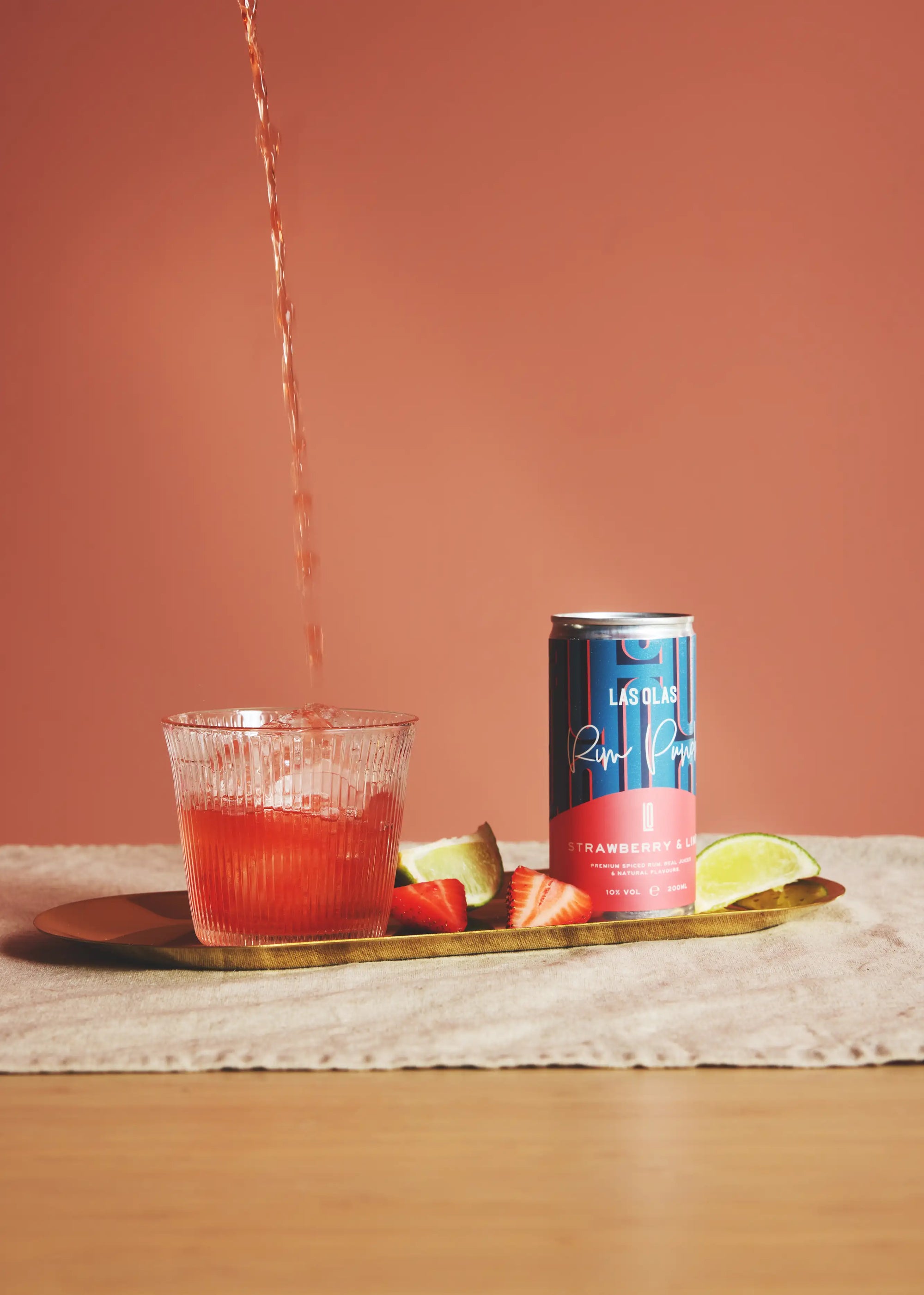las olas strawberry and lime being poured into a glass with a can of 'Las Olas' on a wooden tray against a pink background
