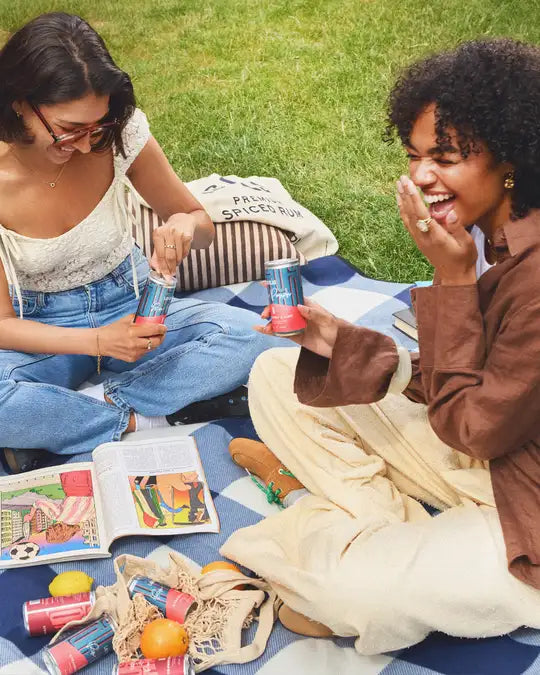 Two women sitting on a blanket outdoors, enjoying las olas rum punch drinks and snacks.